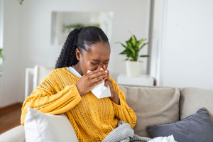 A young woman has a cold and is sneezing. She's sitting on the couch with a tissue to catch her runny nose. She has allergies and is coughing because of them. Signs upholstery furniture needs cleaning concept.