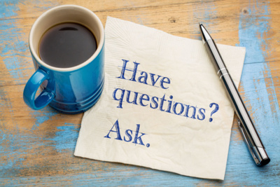 Handwritten note on a napkin with the phrase, "Have Questions? Ask." next to a cup of coffee on a table.